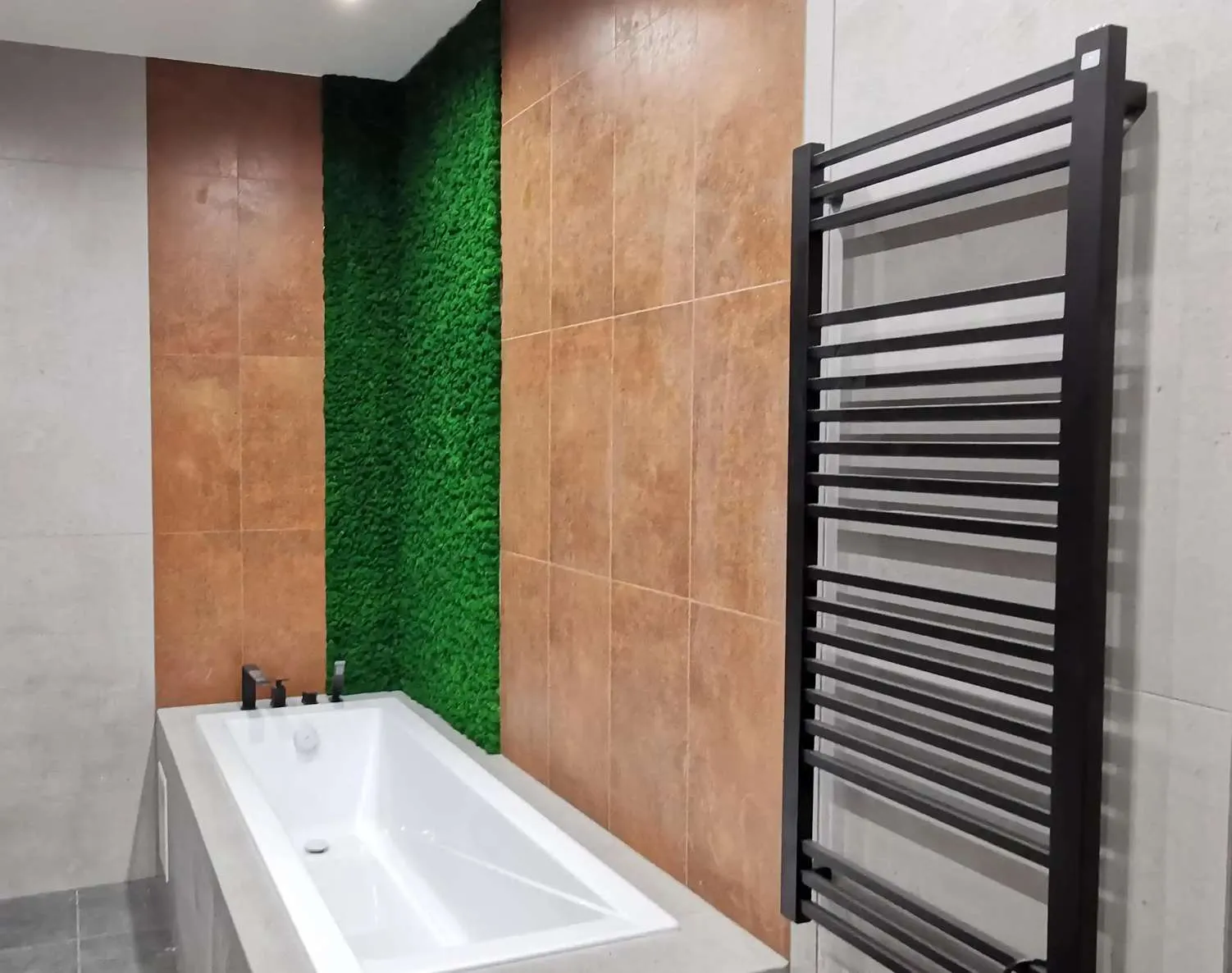 Realizacja mchu stabilizowanego w wąskiej wnęce łazienki obok wbudowanej wanny i czarnego drabinkowego grzejnika. Precyzyjne wykończenie krawędzi.